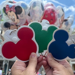 May include: Three Mickey Mouse-shaped keychains with a white background and red, green, and blue accents. The keychains have a white button and a silver ring.
