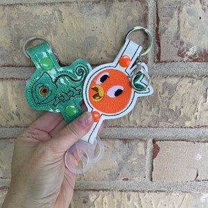 May include: Two keychains with embroidered designs. One keychain features a green chameleon with a white outline. The other keychain features a white and orange fish with a red mouth and black eyes. Both keychains have a white background and a clear plastic ring.