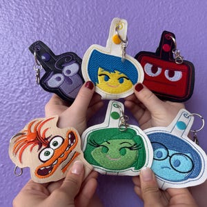 May include: Six embroidered keychains featuring characters from the movie Inside Out. The characters are Joy, Sadness, Anger, Disgust, Fear, and Bing Bong. Each keychain is made of felt and has a metal ring.
