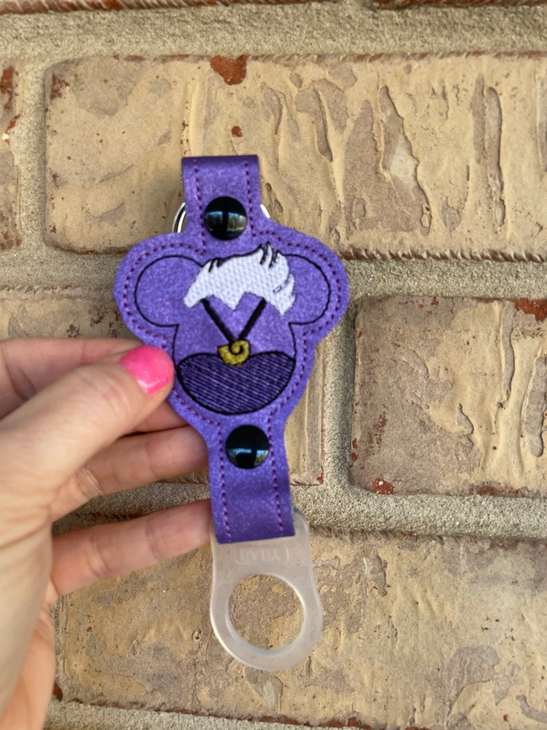 May include: Purple fabric key chain with a cartoon character's head, a white collar, and a gold necklace. The character has white hair and black eyes. The key chain has a clear plastic ring at the bottom.