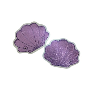 May include: Two purple glitter seashell patches with white stitching. The seashells are sewn onto a white background.
