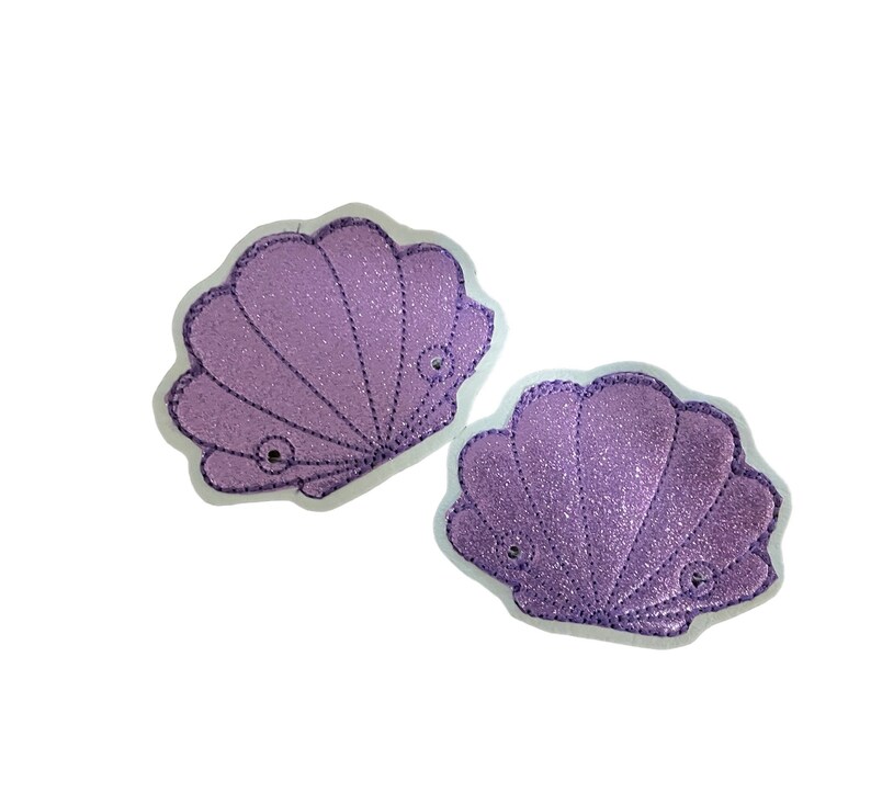 May include: Two purple glitter seashell patches with white stitching. The seashells are sewn onto a white background.