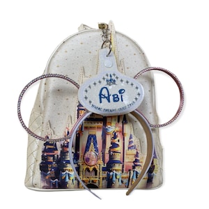 May include: A cream-colored backpack with a castle design and a matching headband with sparkly ears. A personalized name tag with the name "Abi" is attached. The text "WHERE DREAMS COME TRUE" is also visible.