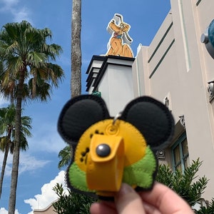 May include: A yellow and green Mickey Mouse plush keychain with black ears. The keychain is in front of a building with a clock tower and a sign featuring a cartoon dog.