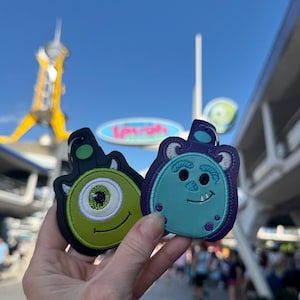 May include: Two embroidered keychains featuring characters from the Disney Pixar movie Monsters Inc. The keychain on the left is green with one large eye and a toothy grin. The keychain on the right is blue with two eyes and a small smile.