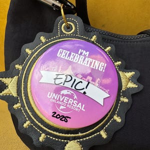May include: A black and gold Universal Orlando Resort tag with a purple and white button that reads "I'm Celebrating! EPIC! Universal Orlando Resort 2025". The tag is attached to a black bag with a silver clasp.