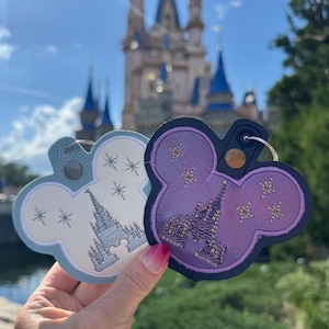 May include: Two Mickey Mouse-shaped keyrings. One is light blue with a white castle design and snowflakes. The other is purple with a gold castle design and stars. Both have key rings and are held up in front of a castle.