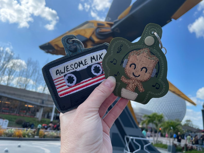 May include: Two embroidered keychains featuring characters from the Guardians of the Galaxy. One keychain is a black cassette tape with the text "Awesome Mix" and the other is a green character with headphones and the text "I am Groot".
