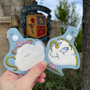 May include: Two embroidered keychains featuring characters from Beauty and the Beast. The keychain on the left is a white teapot with a purple lid and a closed eye. The keychain on the right is a white teacup with a gold handle and an open eye. Both keychains are on a blue background.