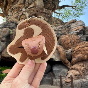 May include: A brown leather keychain shaped like Simba from The Lion King. The keychain has a small pocket for a key or other small item.