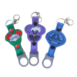 May include: Three colorful keychains with embroidered Disney characters. The keychains feature Ursula, Ariel and Prince Eric, and a green keychain with a seashell and a crown. Each keychain has a metal bottle opener.