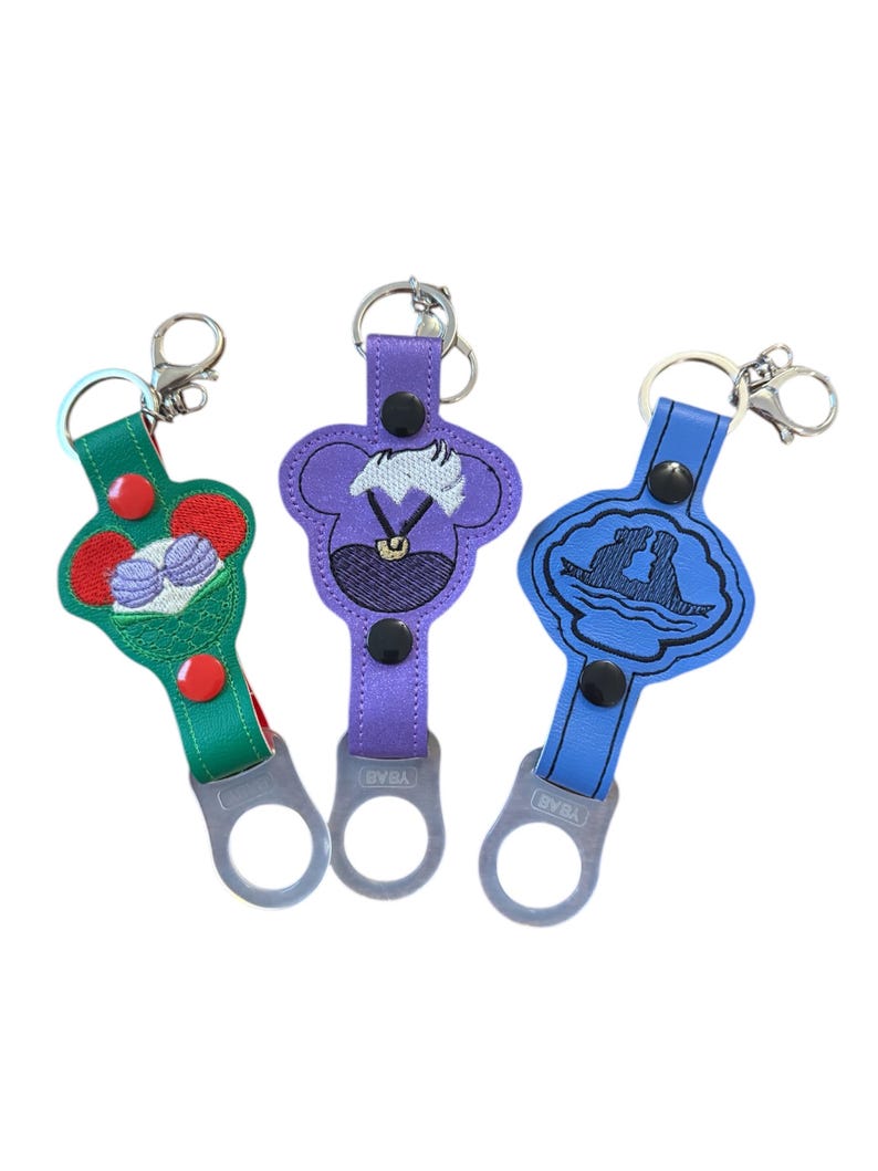 May include: Three colorful keychains with embroidered Disney characters. The keychains feature Ursula, Ariel and Prince Eric, and a green keychain with a seashell and a crown. Each keychain has a metal bottle opener.