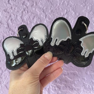 May include: Three black and silver keychains shaped like a dripping ice cream cone with a bow on top. The keychains are made of fabric and have a shiny, iridescent finish.