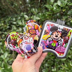 May include: A pair of keychains featuring a colorful candy-themed pattern with a Mickey Mouse-shaped design and a square-shaped holder. The square holder displays a cartoon vampire mouse character surrounded by various sweets and vibrant colors.