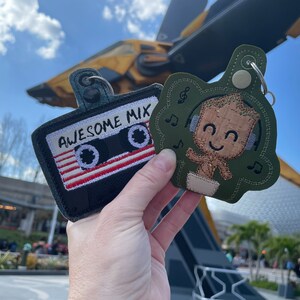 May include: Two embroidered keychains featuring characters from the Guardians of the Galaxy. One keychain is a green and brown Baby Groot wearing headphones with the text "Awesome Mix" on it. The other keychain is a black and red cassette tape.