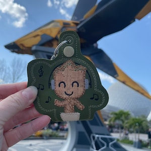 May include: A green felt keychain with a brown embroidered image of a character from the Guardians of the Galaxy movie series. The character is wearing headphones and has a happy expression. The keychain is in front of a large spaceship.