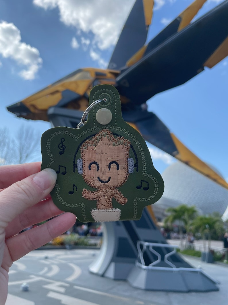 May include: A green felt keychain with a brown embroidered image of a character from the Guardians of the Galaxy movie series. The character is wearing headphones and has a happy expression. The keychain is in front of a large spaceship.