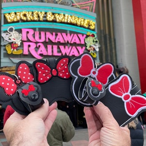 May include: Four black fabric keychains with red and white polka dot bows and red bows. The keychains are shaped like Minnie Mouse ears. The keychains are in front of a sign that says "Mickey & Minnie's Runaway Railway".