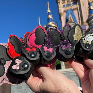 May include: A set of six keychains featuring a black silhouette of Mickey Mouse's head with glitter ears. Each keychain has a different colored bow and glitter on the ears: pink, red, purple, gold, silver, and white.