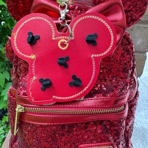 May include: A red sequined backpack featuring a metallic red bow and mouse ears. Attached to the top is a red faux leather pin display board shaped like a mouse head, secured with a silver metal clip. A gold plaque reads Disney Parks Loungefly.