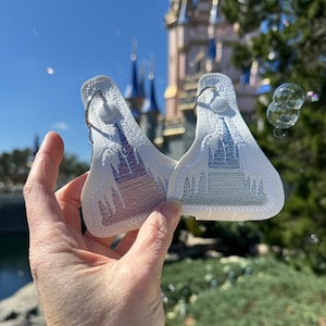 May include: Two white leather keychains with embroidered images of a castle. The castle is in a gradient of blue and purple on one keychain and a gradient of blue and grey on the other keychain.
