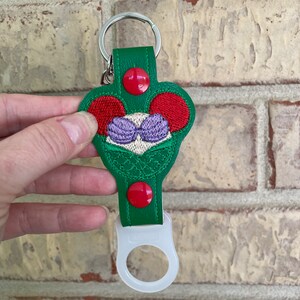 May include: Green leather keychain with a red and white mermaid design. The keychain has a clear plastic bottle opener attached.