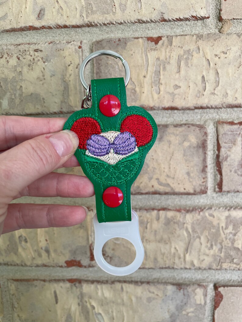 May include: Green leather keychain with a red and white mermaid design. The keychain has a clear plastic bottle opener attached.