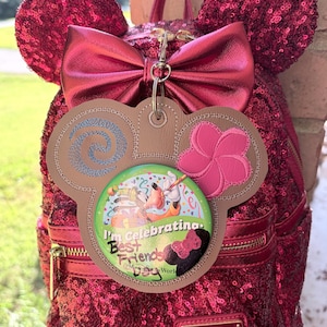 May include: A dark pink sequined backpack with mouse ears and a metallic bow. A tan faux leather tag shaped like mouse ears hangs from the top, featuring a blue spiral, a pink flower, and a button that reads I'm Celebrating Best Friends Day.