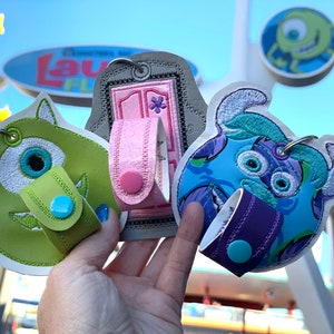May include: Three embroidered keychains featuring characters from the Disney Pixar movie Monsters Inc. The keychains are green, pink, and blue. Each keychain has a small pocket for holding hand sanitiser.
