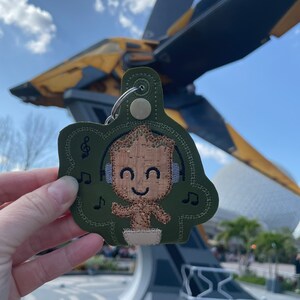 May include: A green fabric keychain with an embroidered image of a character from the Guardians of the Galaxy movie. The character is wearing headphones and has a happy expression. The keychain is in front of a large spaceship.