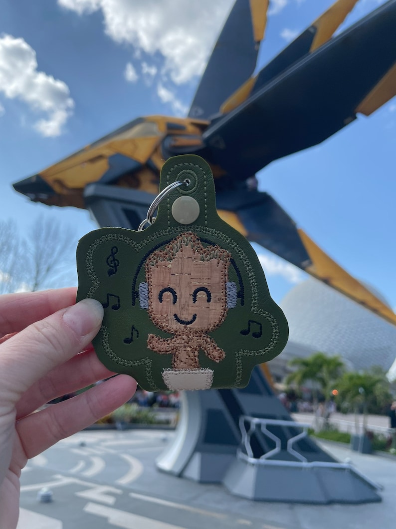 May include: A green fabric keychain with an embroidered image of a character from the Guardians of the Galaxy movie. The character is wearing headphones and has a happy expression. The keychain is in front of a large spaceship.