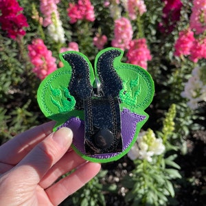 May include: A green, purple, and black embroidered keychain featuring a stylized design. The keychain has a green base with black horns and a purple accent. A silver metal clasp is attached to the top.