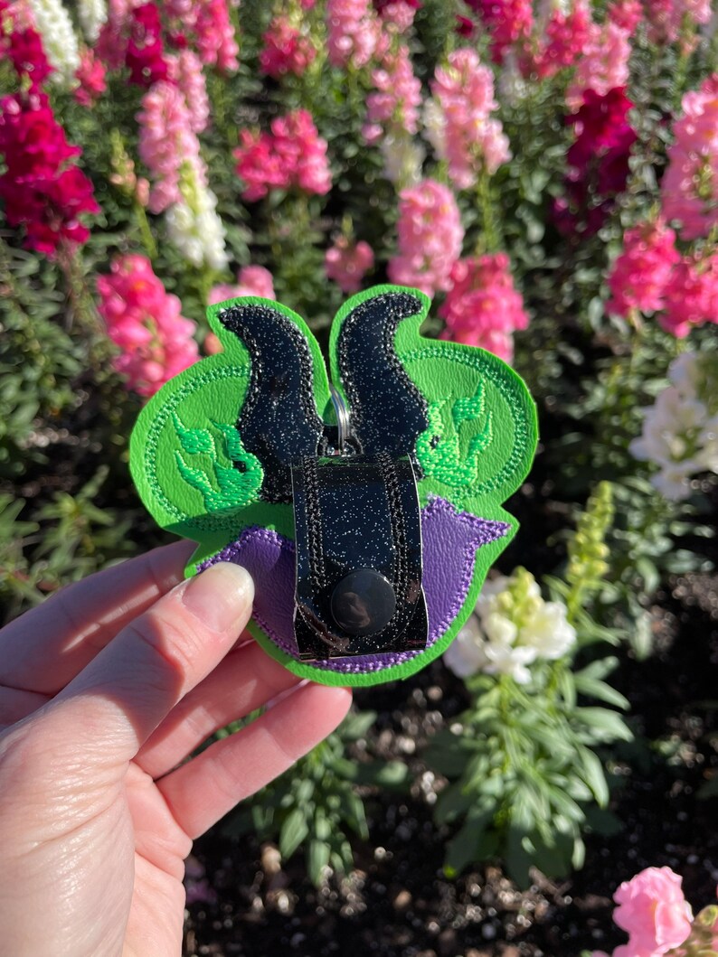May include: A green, purple, and black embroidered keychain featuring a stylized design. The keychain has a green base with black horns and a purple accent. A silver metal clasp is attached to the top.