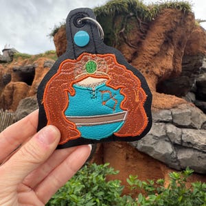 May include: A black leather keychain with a colorful embroidered image of a cartoon character with red hair, a blue dress, and a green crown. The character is holding a blue bucket with a brown handle.