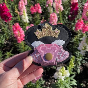 May include: A black, pink, and gold keychain featuring a crown design. The keychain has a gold crown, pink bodice, and a black outline. The background is a blurred view of pink and white flowers.