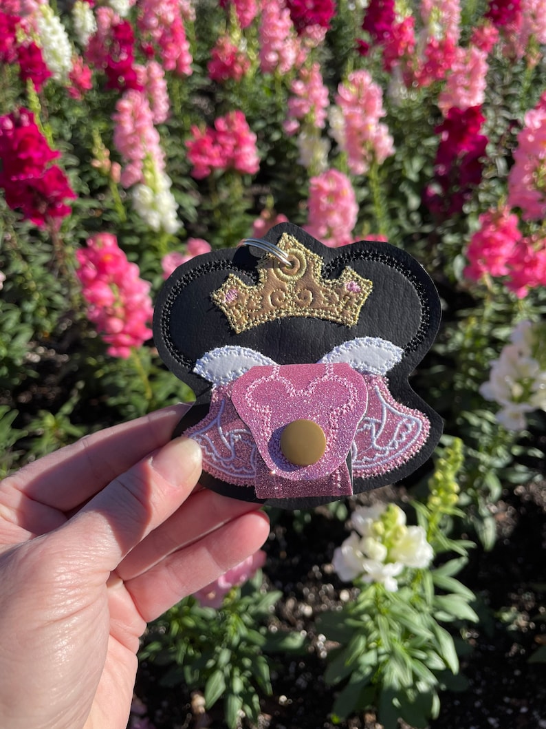 May include: A black, pink, and gold keychain featuring a crown design. The keychain has a gold crown, pink bodice, and a black outline. The background is a blurred view of pink and white flowers.