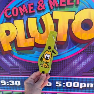 May include: Green fabric Pluto character keychain with a yellow and brown embroidered image of Pluto. The keychain is attached to a sign that says "Come & Meet Pluto".