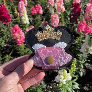 May include: A black, pink, and gold keychain featuring a crown design. The keychain has a pink heart-shaped pocket with a gold button closure. The background is a blurred view of pink and white flowers.