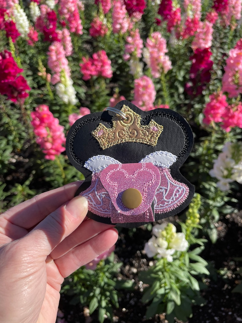 May include: A black, pink, and gold keychain featuring a crown design. The keychain has a pink heart-shaped pocket with a gold button closure. The background is a blurred view of pink and white flowers.