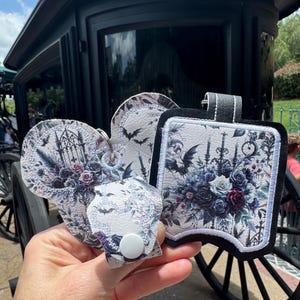 May include: A collection of gothic-themed accessories, including a luggage tag and keychains. The designs feature floral arrangements, bats, and architectural elements in shades of purple, gray, and black. The items are held in front of a black carriage.