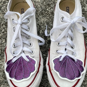 May include: A pair of white canvas sneakers with purple glitter seashell patches on the toes. The laces are white and the shoes have red trim around the soles.