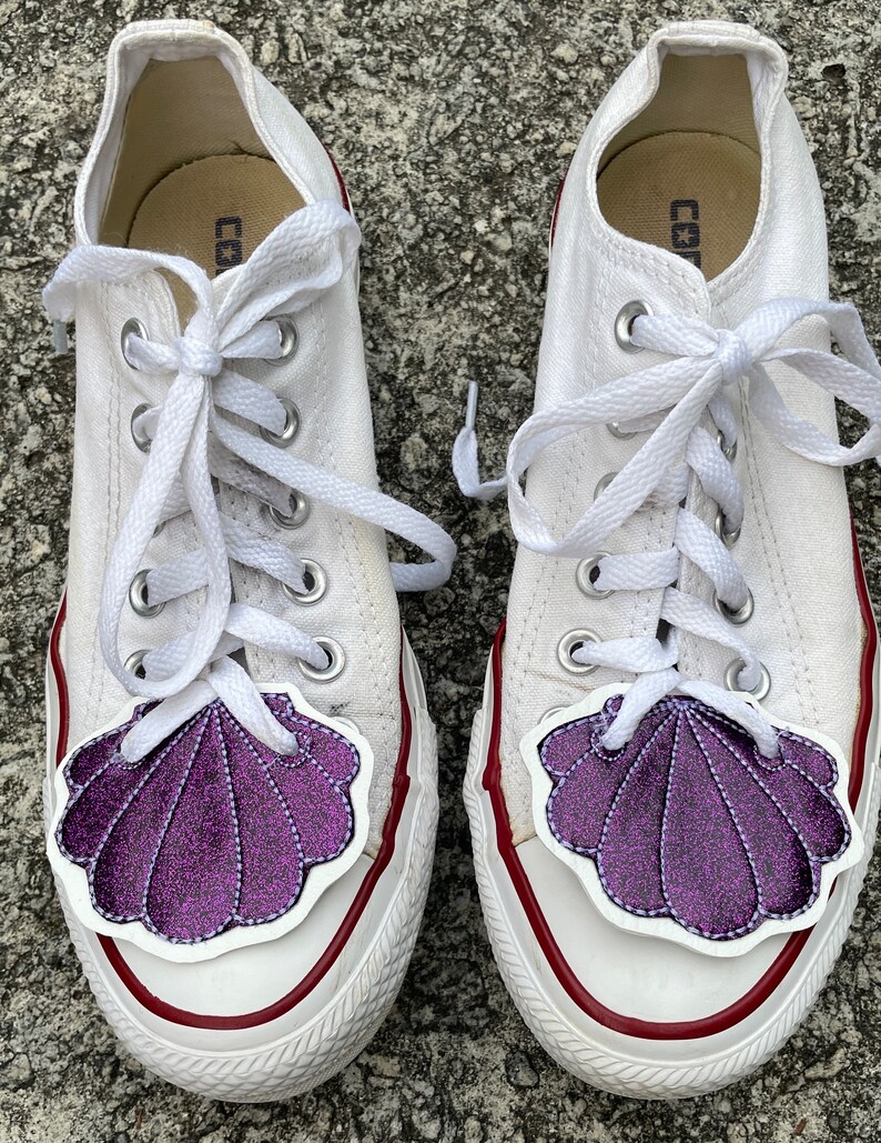 May include: A pair of white canvas sneakers with purple glitter seashell patches on the toes. The laces are white and the shoes have red trim around the soles.