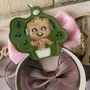 May include: A green felt character keychain with a brown and tan character wearing headphones and a diaper. The character is surrounded by musical notes. The keychain is attached to a pink and white headband with a brown bow.