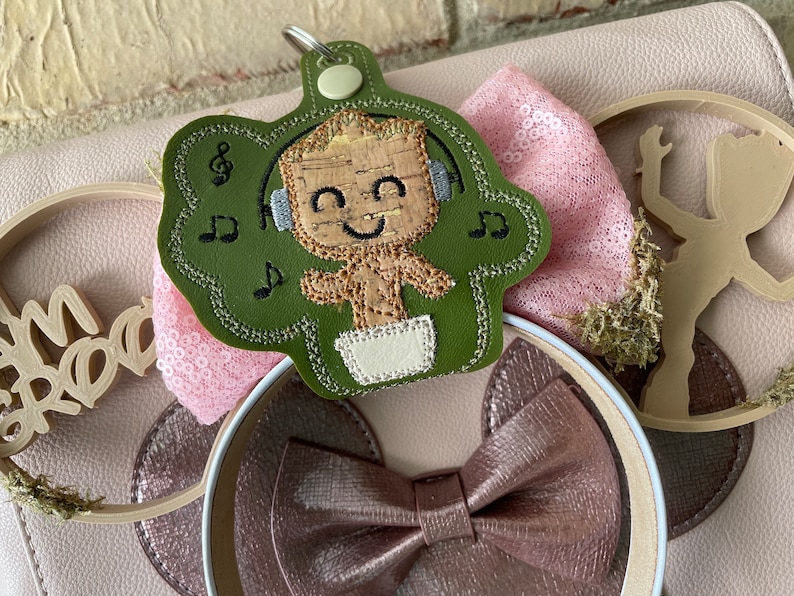 May include: A green felt character keychain with a brown and tan character wearing headphones and a diaper. The character is surrounded by musical notes. The keychain is attached to a pink and white headband with a brown bow.