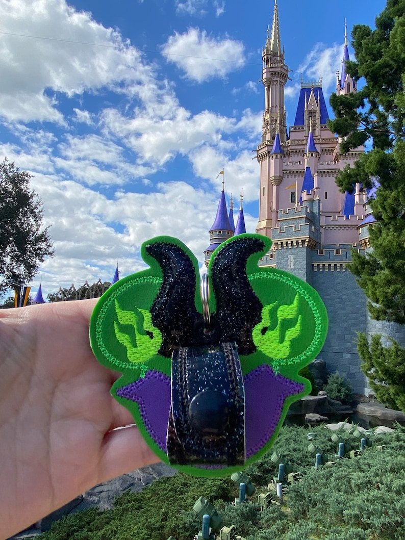 May include: A green, purple, and black patch featuring a stylized design with a black glittered element. The patch is held in front of a castle with blue turrets under a blue sky.