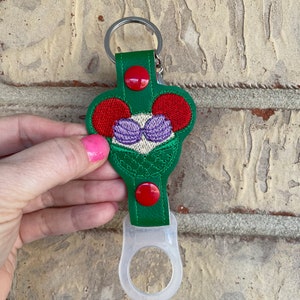May include: Green fabric bottle holder with a red button and a white silicone ring. The fabric has an embroidered design of a mermaid with red hair and purple seashells.