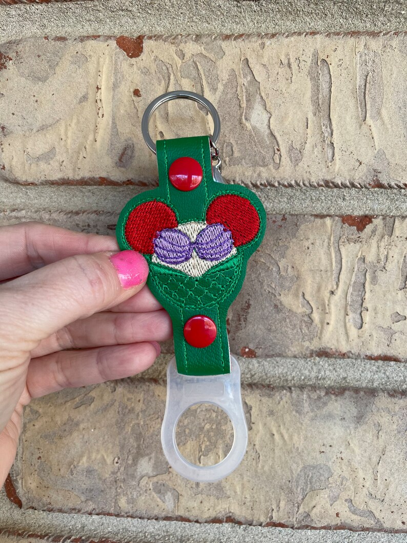 May include: Green fabric bottle holder with a red button and a white silicone ring. The fabric has an embroidered design of a mermaid with red hair and purple seashells.