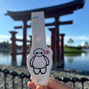 May include: White fabric keychain featuring a black embroidered image of Baymax from the Disney film Big Hero 6. Baymax is holding a pink speech bubble.