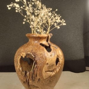 May include: A natural wood vase with a polished finish and organic, irregular openings in the body. The vase features a rounded shape with a narrow neck and is filled with a cluster of delicate, dried white baby's breath flowers.