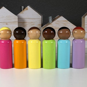 May include: Seven wooden peg dolls painted in rainbow colors, standing in a row on a white surface. The dolls have brown hair and painted faces.